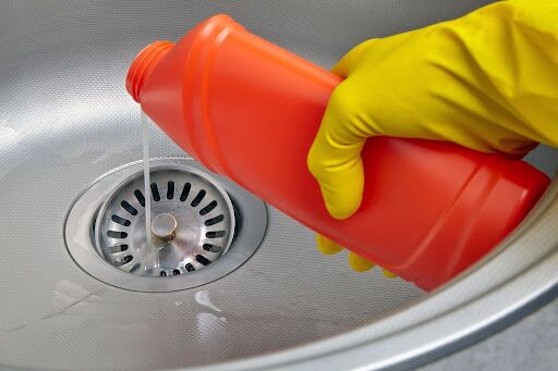 A gloved hand pouring chemical drain cleaner down a sink drain.